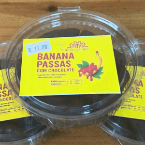 Pote transparente redondo contendo bananas passas banhadas em chocolate meio amargo, com rótulo amarelo vibrante da marca Sítio do Campo