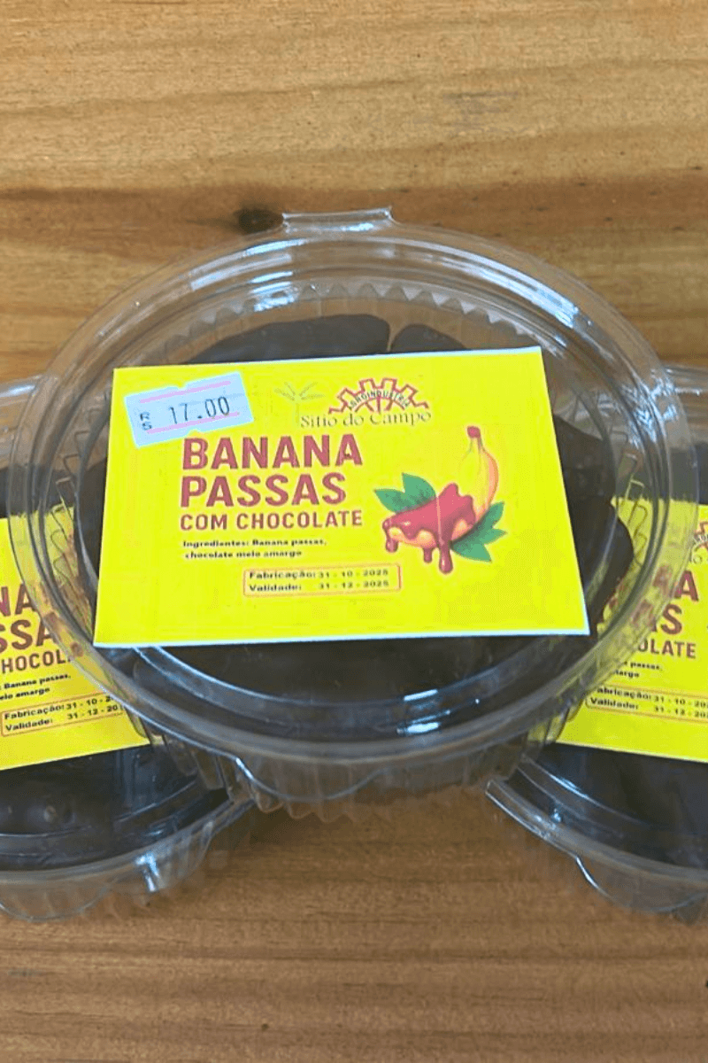 Pote transparente redondo contendo bananas passas banhadas em chocolate meio amargo, com rótulo amarelo vibrante da marca Sítio do Campo