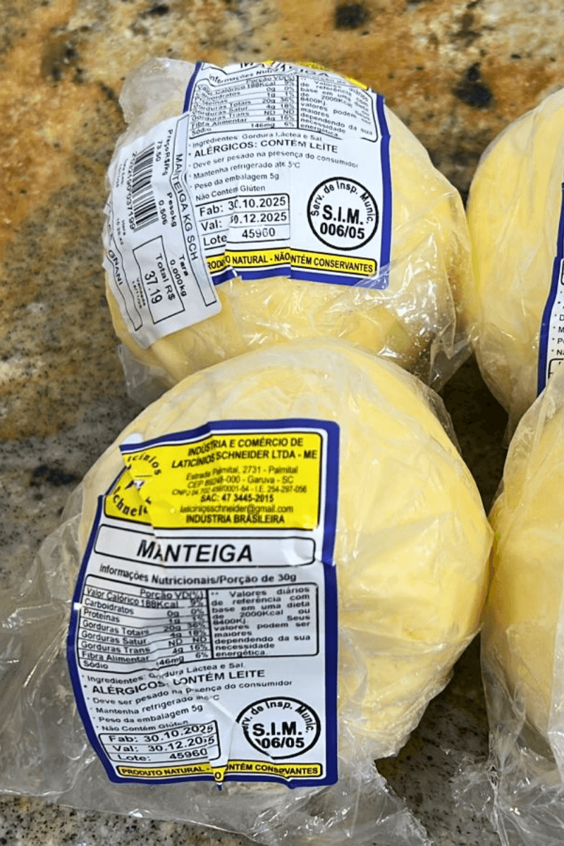 Três unidades de Manteiga Bola artesanal da Laticínios Schneider embaladas em plástico transparente sobre balcão, destacando o rótulo amarelo e selo de inspeção.