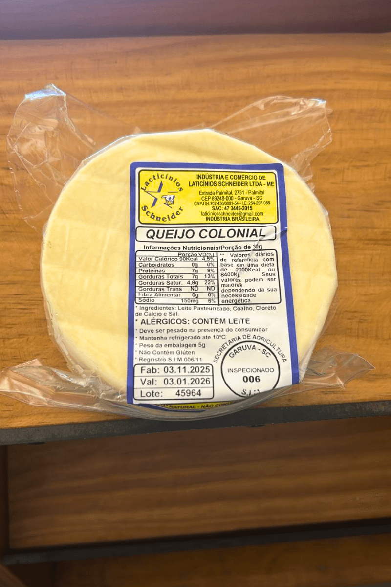 Queijo Colonial artesanal da Laticínios Schneider, peça inteira embalada a vácuo sobre superfície de madeira, evidenciando a textura clara e o selo de inspeção.