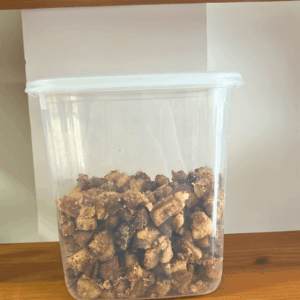 Pote plástico transparente repleto de torresmo artesanal crocante em pedaços, organizado em uma prateleira de madeira rústica.