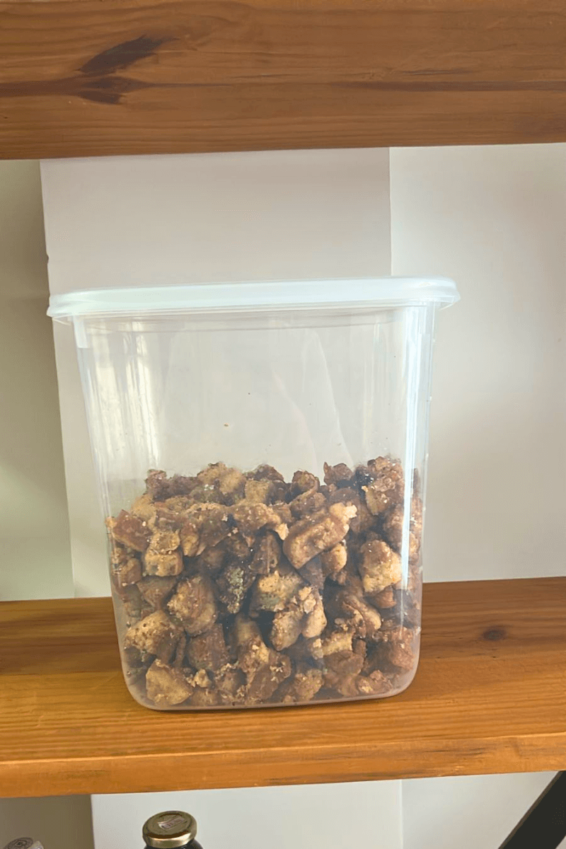 Pote plástico transparente repleto de torresmo artesanal crocante em pedaços, organizado em uma prateleira de madeira rústica.