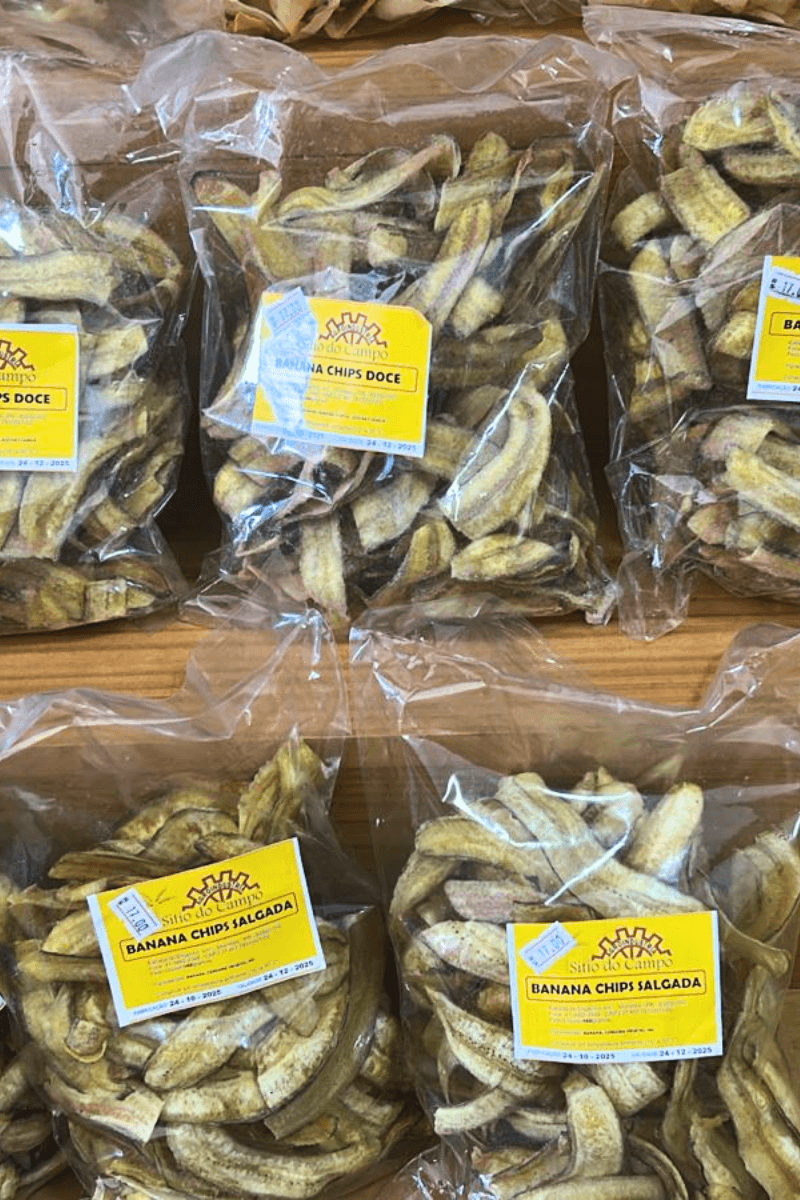 Vários pacotes de Banana Chips Sítio do Campo expostos em uma prateleira, mostrando as versões Doce e Salgada com rótulos amarelos.