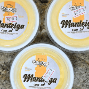 Três potes redondos de Manteiga com Sal da marca Melkland vistos de cima, apresentando rótulo branco e amarelo com a frase "nós amamos queijo" e indicação de produção em Castro, Paraná.