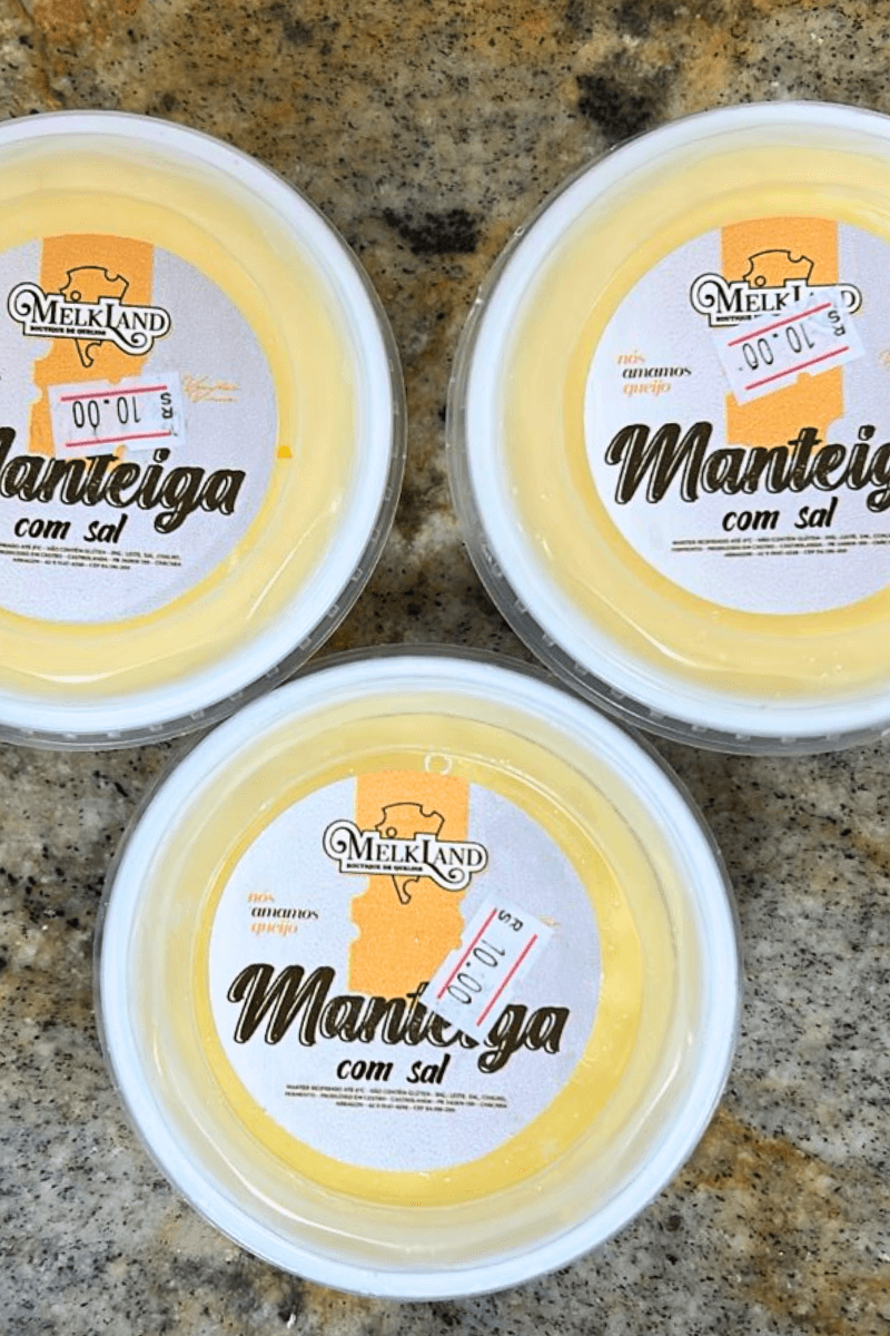 Três potes redondos de Manteiga com Sal da marca Melkland vistos de cima, apresentando rótulo branco e amarelo com a frase "nós amamos queijo" e indicação de produção em Castro, Paraná.