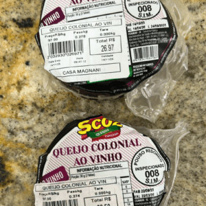 Duas peças fracionadas de Queijo Colonial ao Vinho da marca Scoz, embaladas a vácuo, com casca de tonalidade arroxeada e rótulo destacando o selo de inspeção S.I.M. 008 de Pouso Redondo.