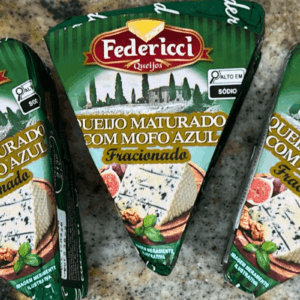 Três peças triangulares de Queijo Maturado com Mofo Azul da marca Federicci, embaladas em papel verde com ilustração de figos e nozes, destacando o rótulo "Alto em Sódio" e o selo de inspeção.