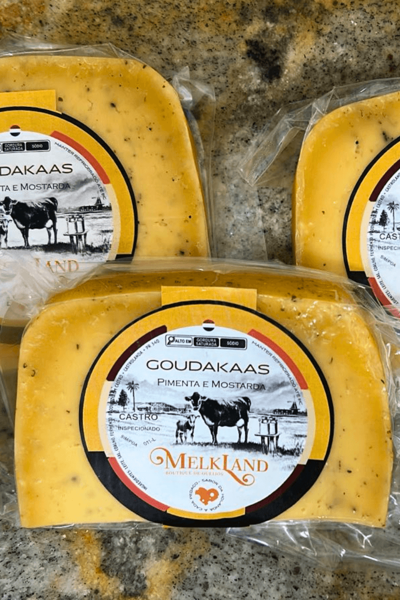 Peças de Queijo Goudakaas com Pimenta e Mostarda da marca Melkland, embaladas a vácuo, mostrando a massa amarela com pontos de especiarias e rótulo circular amarelo e preto.